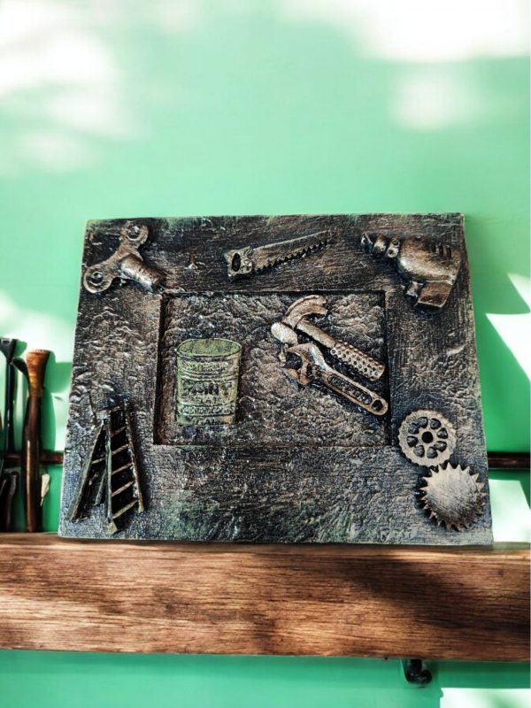 a small , three-dimensional artwork featuring miniature metal tools and objects, including a saw, drill, hammer, wrench, barrel, ladder and gears, arranged on a rectangular base.