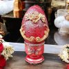 Handcrafted red decorative egg with gold floral details on a matching stand, set against a floral backdrop.
